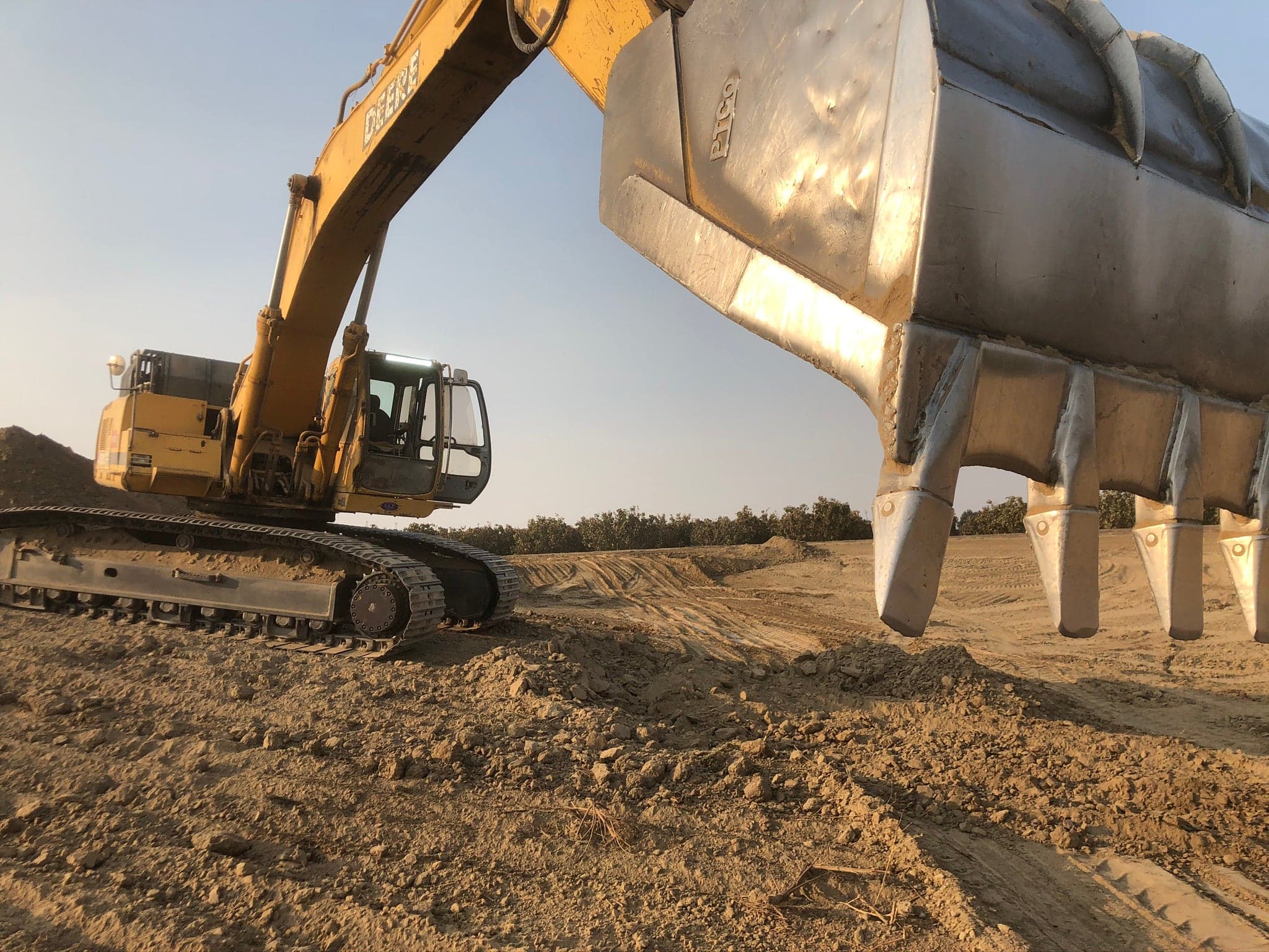 A John Deere excavator with a large toothed bucket working graded ground alongside an orchard