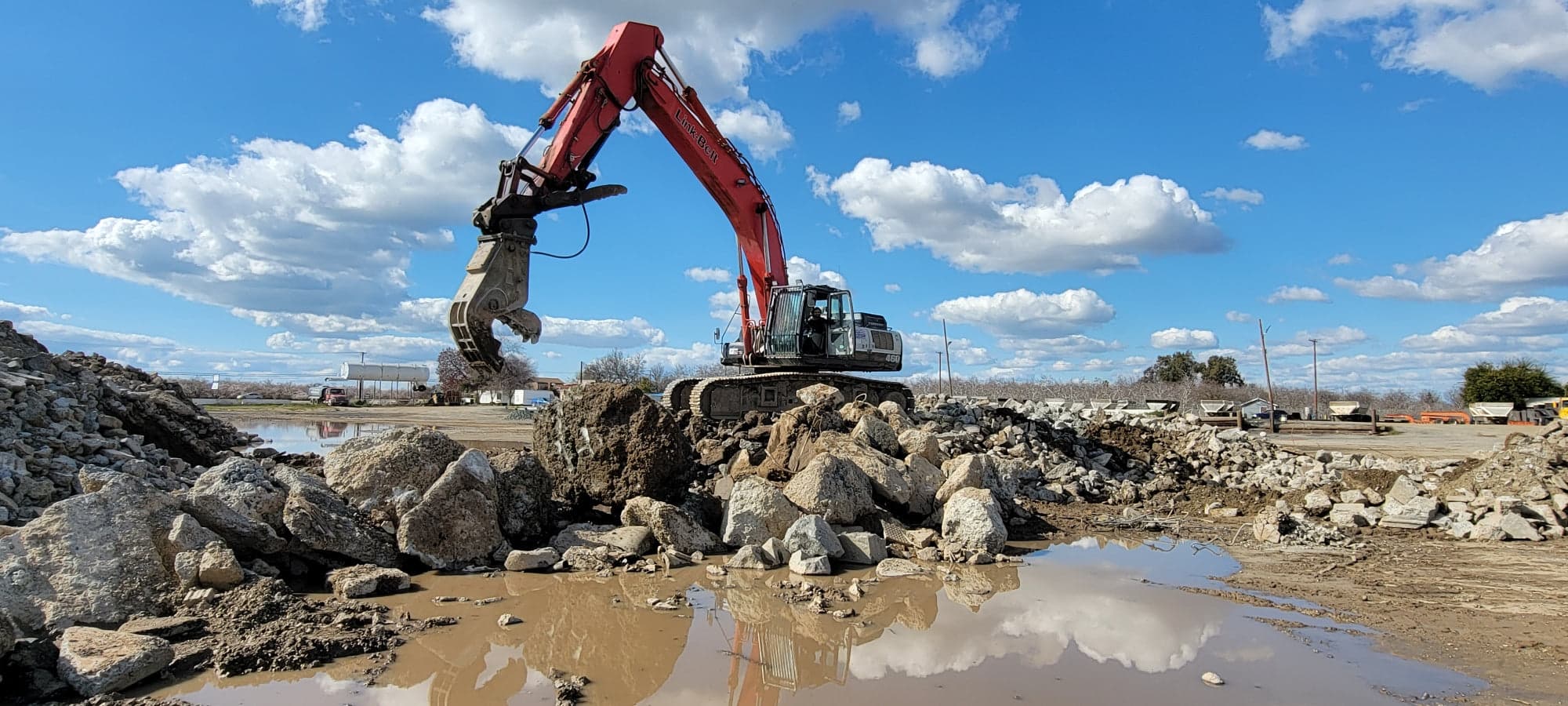 An excavator fitted with a concrete pulverizer standing on a pile of broken demolition concrete at a Central Valley job site